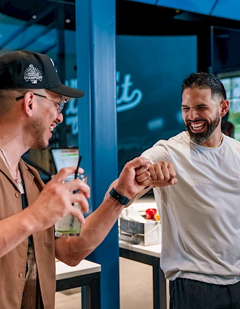 Two men are smiling and bumping fists in a casual indoor setting.
